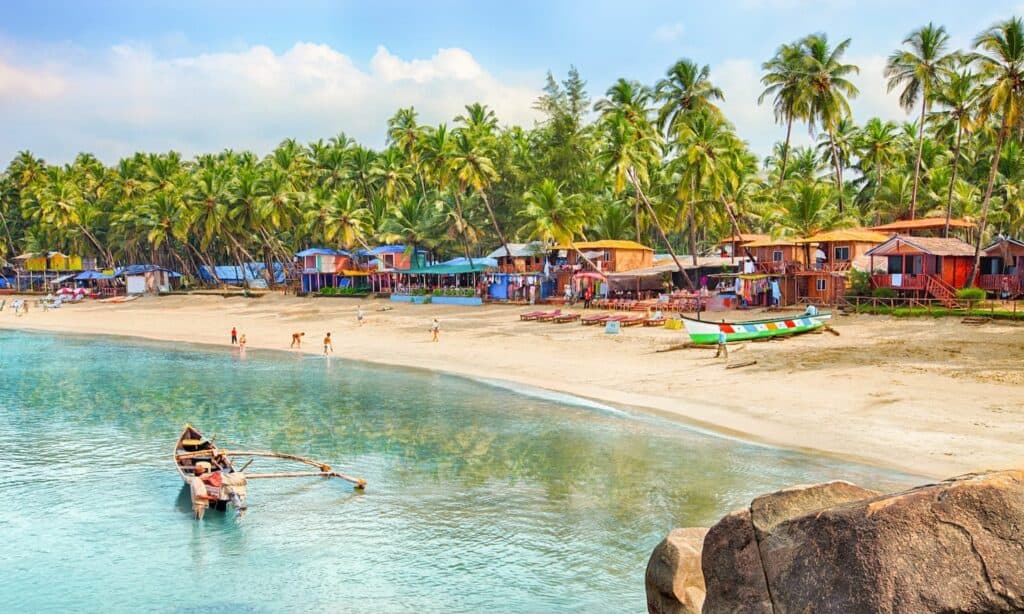 Tropical beach scene in Goa with colorful beach huts, palm trees, fishing boats near the shore, and tourists walking along the clear sandy shoreline under a partly cloudy sky