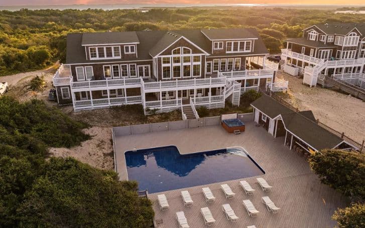 Luxstay USA's luxury Outer Banks vacation home featuring a large pool, multiple white lounge chairs on a spacious deck, a hot tub, and adjacent buildings surrounded by greenery at sunset.